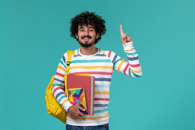 Vista frontal do estudante do sexo masculino usando mochila amarela segurando arquivos e o caderno na parede azul