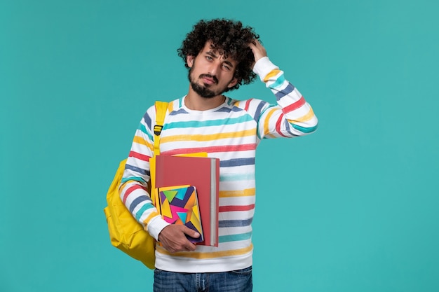 Vista frontal do estudante do sexo masculino usando mochila amarela segurando arquivos e o caderno na parede azul