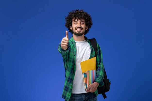 Vista frontal do estudante do sexo masculino usando uma mochila preta segurando cadernos e arquivos sorrindo na parede azul