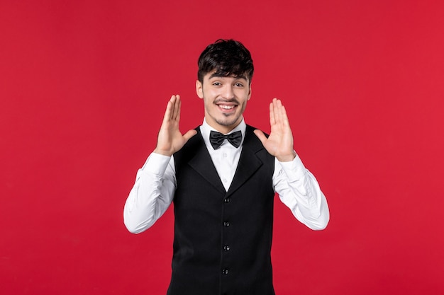 Vista frontal do garçom feliz e satisfeito em um uniforme com uma borboleta no pescoço e fundo vermelho