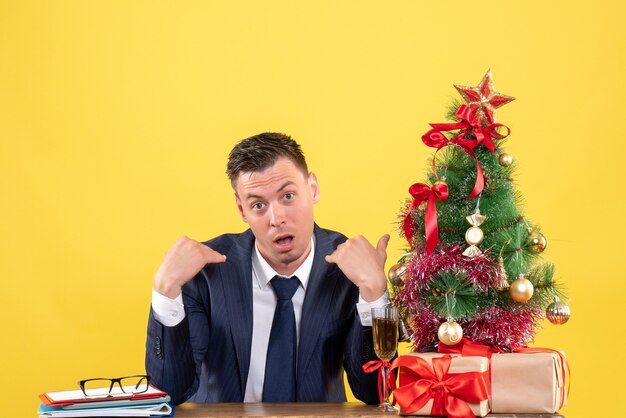 Vista frontal do homem admirado, apontando para si mesmo sentado à mesa perto da árvore de natal e presentes em amarelo