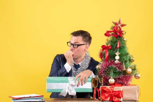 Vista frontal do homem admirado, olhando para algo sentado à mesa perto da árvore de natal e presentes em amarelo