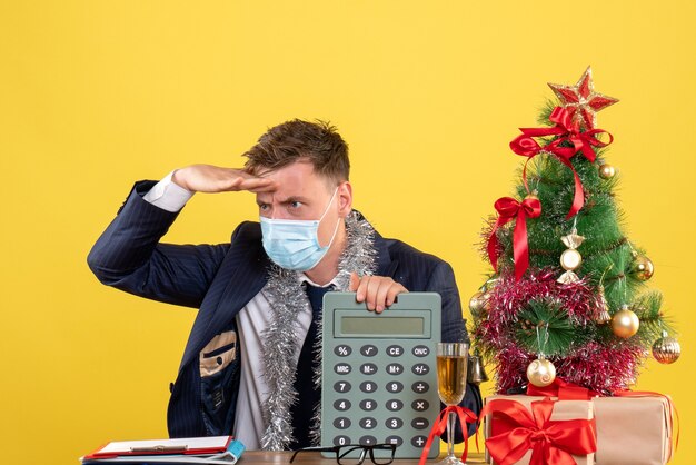 Vista frontal do homem de negócios colocando a mão na testa, sentado à mesa perto da árvore de natal e presentes em amarelo