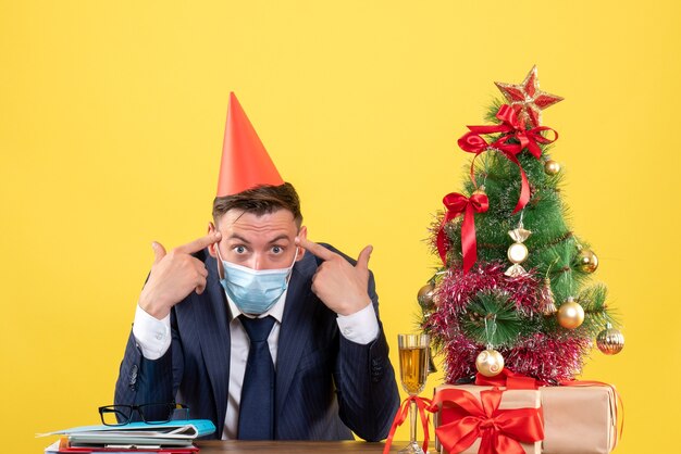 Vista frontal do homem de negócios colocando os dedos em sua têmpora, sentado à mesa perto da árvore de natal e presentes em amarelo