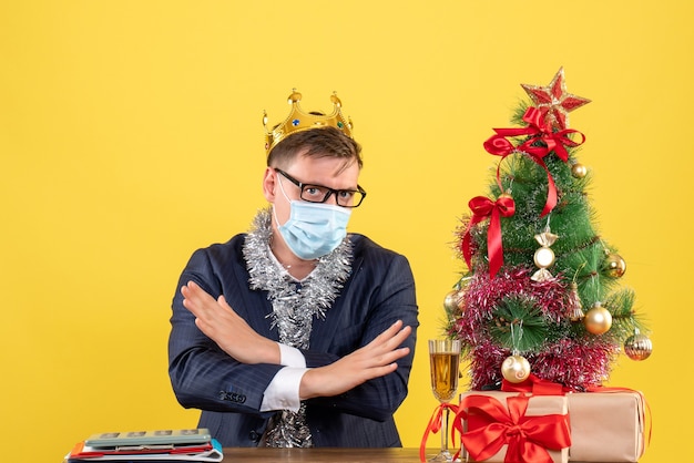 Vista frontal do homem de negócios cruzando as mãos, sentado à mesa perto da árvore de natal e presentes na parede amarela