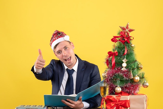 Vista frontal do homem de negócios fazendo sinal de polegar para cima sentado à mesa perto da árvore de natal e presentes em amarelo