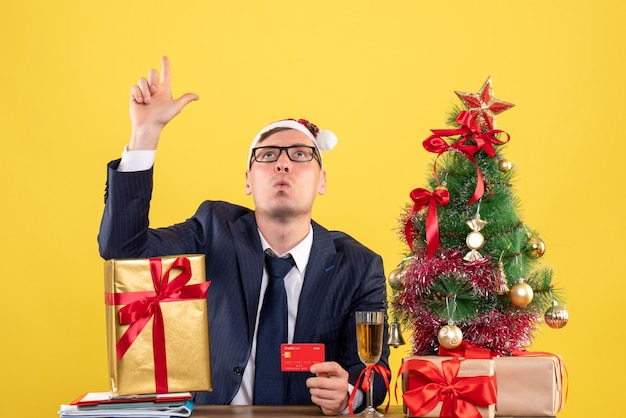 Foto grátis vista frontal do homem de negócios olhando sentado à mesa perto da árvore de natal e presentes em amarelo