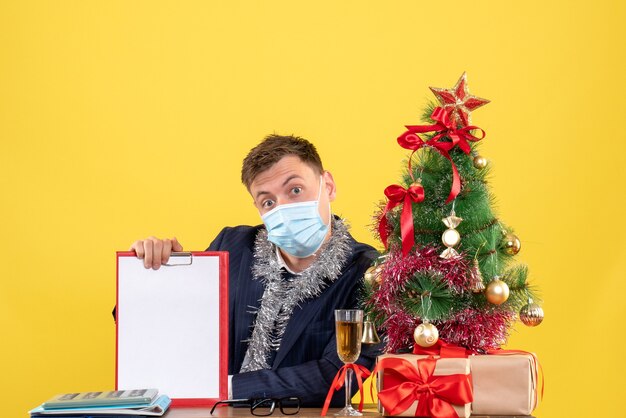 Vista frontal do homem de negócios segurando a prancheta, sentado à mesa perto da árvore de natal e presentes em amarelo