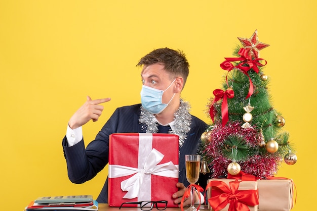 Vista frontal do homem de negócios segurando seu presente sentado à mesa perto da árvore de natal e presentes em amarelo