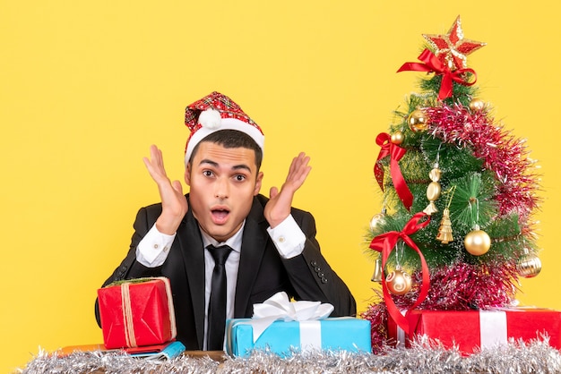 Vista frontal do homem de terno com chapéu de papai noel sentado na mesa árvore de natal e presentes