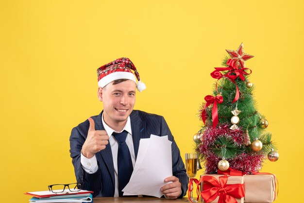 Vista frontal do homem satisfeito fazendo sinal de polegar para cima sentado à mesa perto da árvore de natal e presentes em amarelo