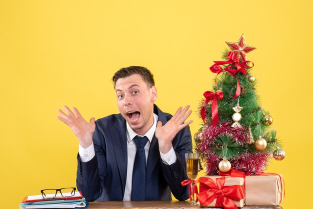 Vista frontal do homem surpreso com as mãos abertas, abrindo a boca, sentado à mesa perto da árvore de natal e presentes