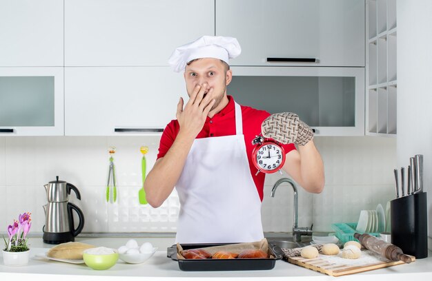 Vista frontal do jovem chef masculino surpreso usando o suporte segurando o relógio na cozinha branca