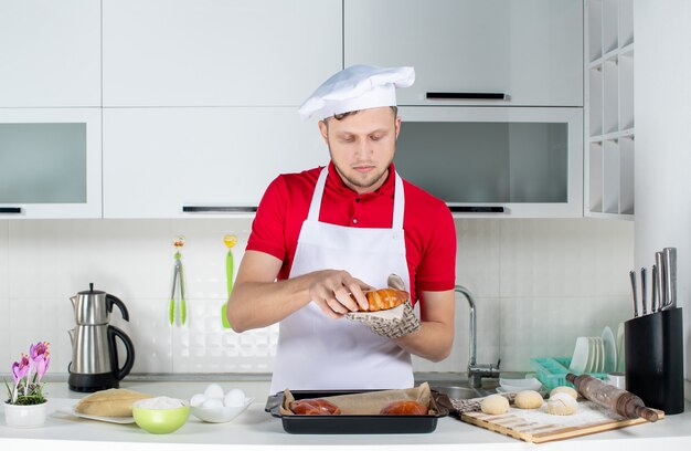Vista frontal do jovem chef ocupado usando um suporte segurando um dos doces recém-assados na cozinha branca