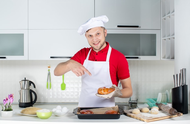 Vista frontal do jovem chef sorridente usando o suporte, segurando e apontando um dos doces recém-assados na cozinha branca