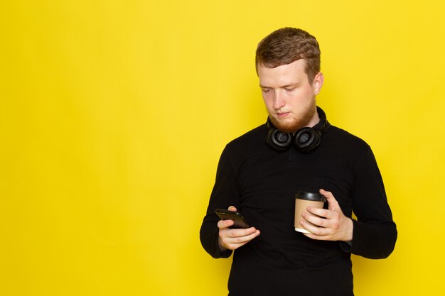 Vista frontal do jovem de camisa preta, usando telefone e segurando a xícara de café