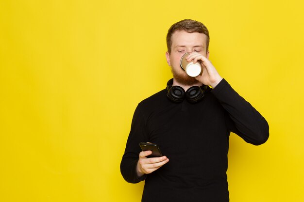 Vista frontal do jovem de camisa preta, usando telefone e tomando café