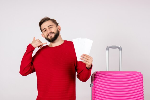 Vista frontal do jovem homem com bolsa rosa e segurando os bilhetes na parede branca, viagem, viagem, voo, férias, turista