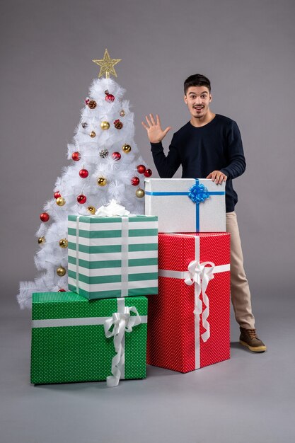 Vista frontal do jovem macho com presentes e árvore de natal no piso cinza presente de natal ano novo