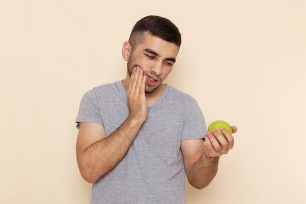 Foto grátis vista frontal do jovem macho em uma camiseta cinza com dor de dente segurando uma maçã verde em bege