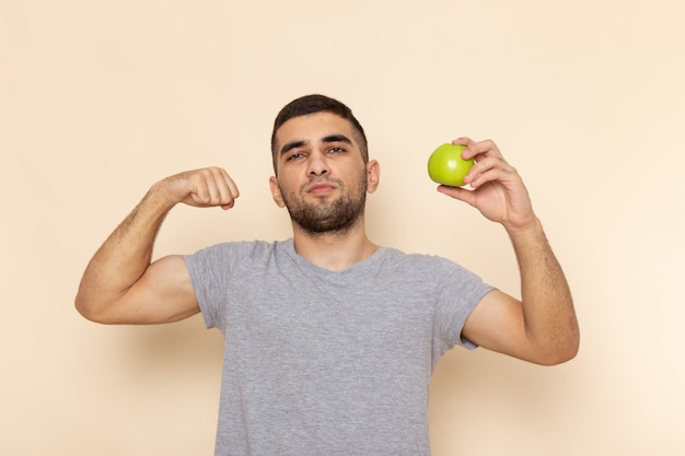 Vista frontal do jovem macho em uma camiseta cinza segurando uma maçã e flexionando em bege