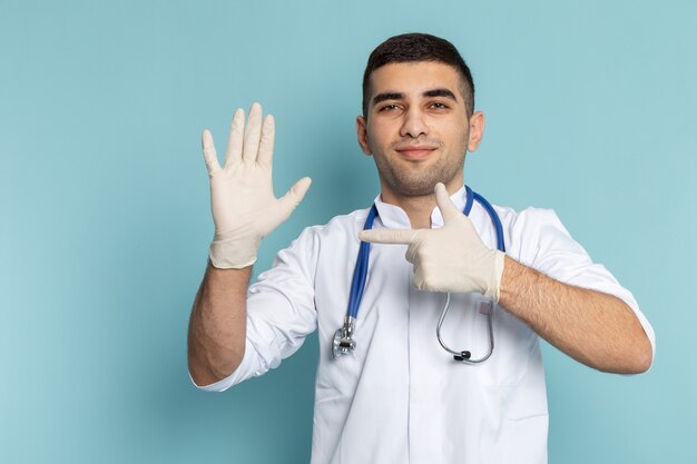 Vista frontal do jovem médico de terno branco com estetoscópio azul apontando para a luva