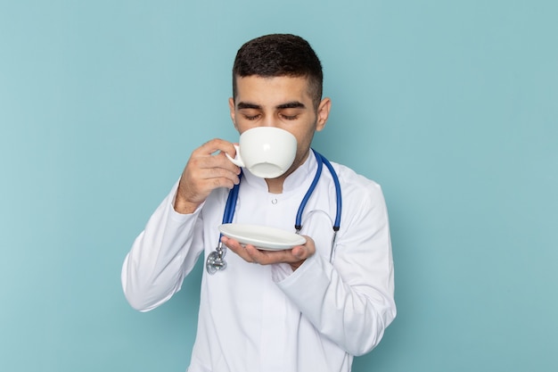 Vista frontal do jovem médico de terno branco com estetoscópio azul bebendo café