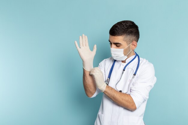 Vista frontal do jovem médico de terno branco com estetoscópio azul e sorrindo usando luvas