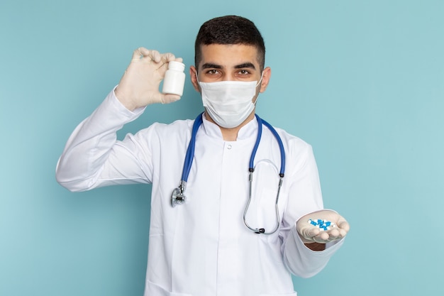 Vista frontal do jovem médico de terno branco com estetoscópio azul segurando comprimidos
