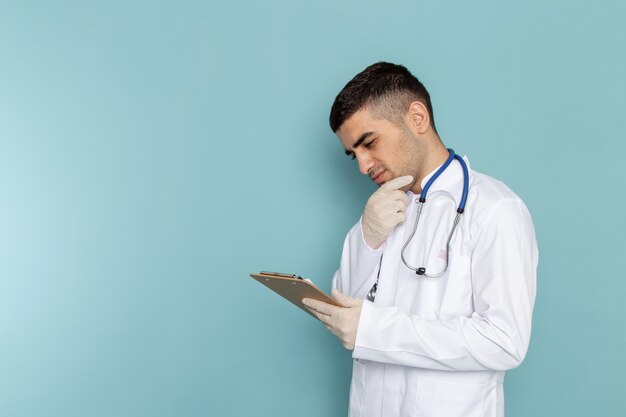 Vista frontal do jovem médico de terno branco com estetoscópio azul segurando o bloco de notas com expressão pensativa