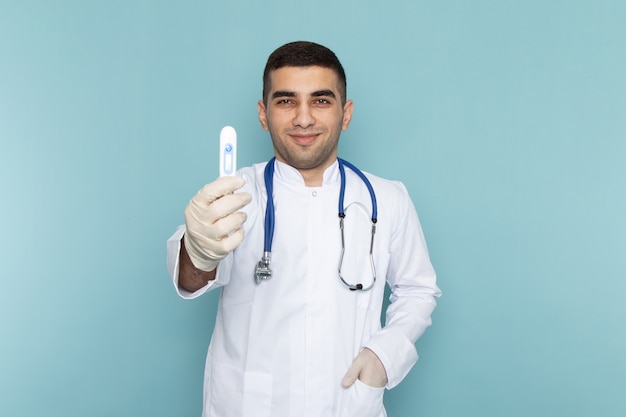 Vista frontal do jovem médico de terno branco com estetoscópio azul sorrindo e segurando o dispositivo