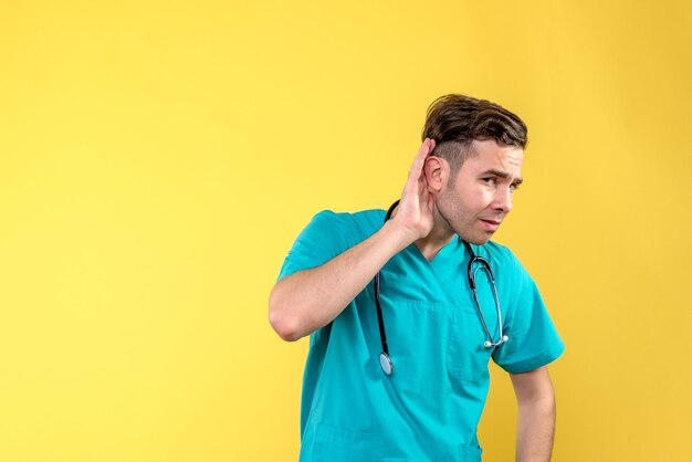 Vista frontal do jovem médico ouvindo na parede amarela