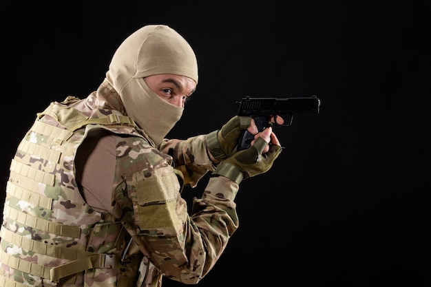 Vista frontal do jovem soldado de uniforme apontando a arma na parede preta