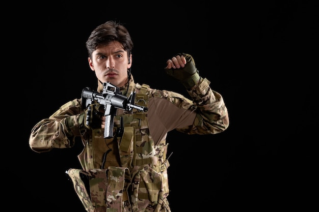 Vista frontal do jovem soldado de uniforme com rifle na parede preta