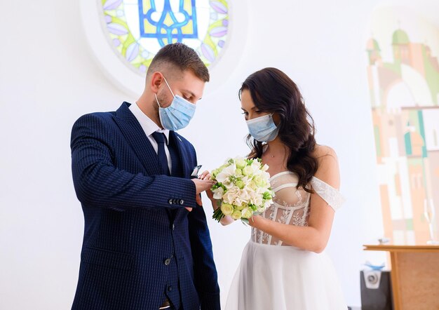 Vista frontal do lindo casal que usava roupas de casamento e máscaras protetoras nos rostos trocando por anéis durante o casamento civil dentro do cartório