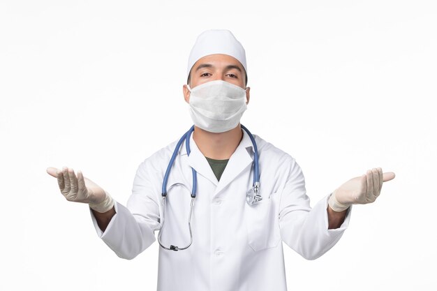 Vista frontal do médico do sexo masculino em traje médico e máscara devido à doença do vírus pandêmico do covid-vírus da parede branca-clara