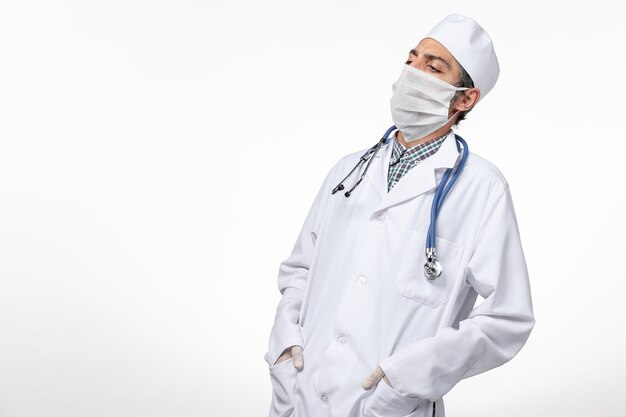 Vista frontal do médico em terno branco e máscara devido a coronavírus posando na mesa branca