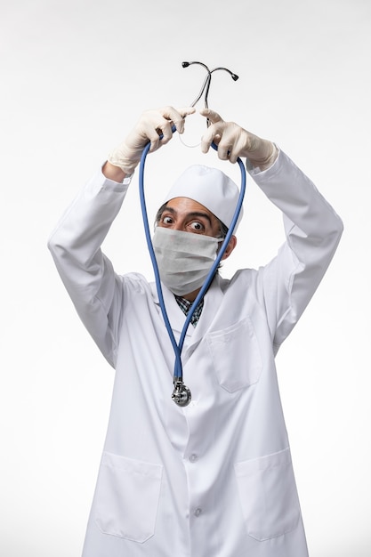 Vista frontal do médico masculino com traje e máscara devido a coronavírus segurando um estetoscópio azul na superfície branca