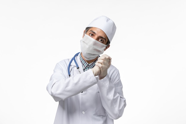 Vista frontal do médico masculino com uniforme e máscara devido a coronavírus na superfície branca