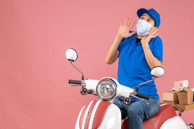 Vista frontal do mensageiro com máscara médica usando chapéu, sentado na scooter e olhando para cima com uma expressão facial de medo no fundo cor de pêssego