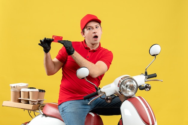Vista frontal do mensageiro curioso usando blusa vermelha e luvas de chapéu na máscara médica, entregando pedidos sentado na scooter, mostrando o cartão do banco