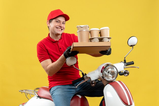 Vista frontal do mensageiro feliz sorridente, vestindo uma blusa vermelha e luvas de chapéu na máscara médica, entregando pedidos sentado na scooter segurando pedidos