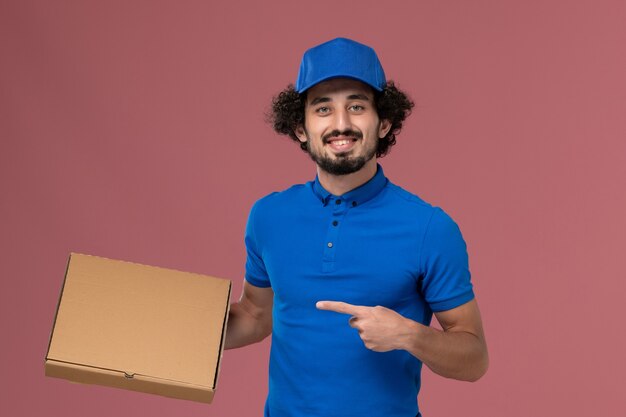 Vista frontal do mensageiro masculino com boné de uniforme azul e caixa de comida nas mãos na parede rosa claro