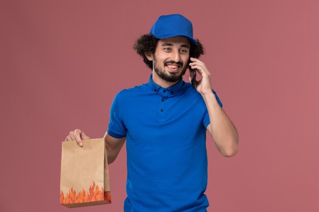 Vista frontal do mensageiro masculino com boné de uniforme azul e entrega de pacote de comida nas mãos, falando ao telefone na parede rosa