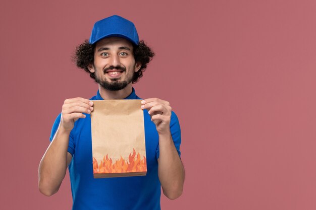 Foto grátis vista frontal do mensageiro masculino com boné uniforme azul e pacote de comida de papel nas mãos na parede rosa claro