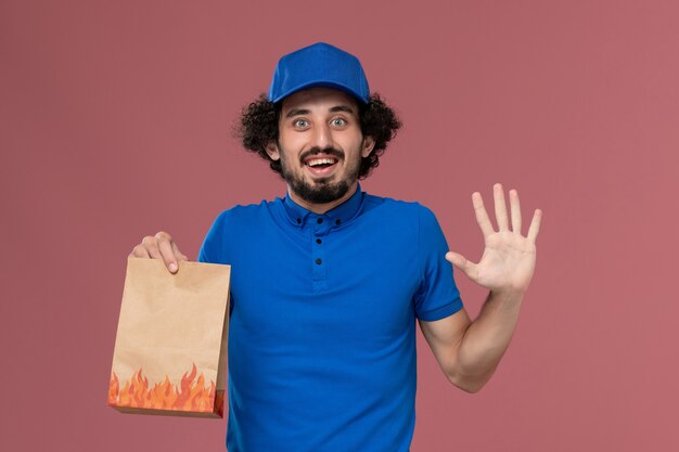 Vista frontal do mensageiro masculino com boné uniforme azul e pacote de comida de papel nas mãos na parede rosa claro