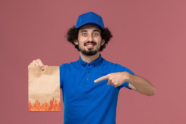 Vista frontal do mensageiro masculino com boné uniforme azul e pacote de comida de papel nas mãos na parede rosa claro