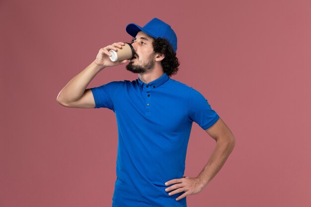 Vista frontal do mensageiro masculino com uniforme azul e boné bebendo café na parede rosa