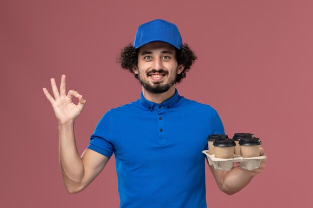 Vista frontal do mensageiro masculino com uniforme azul e boné com xícaras de café nas mãos na parede rosa claro