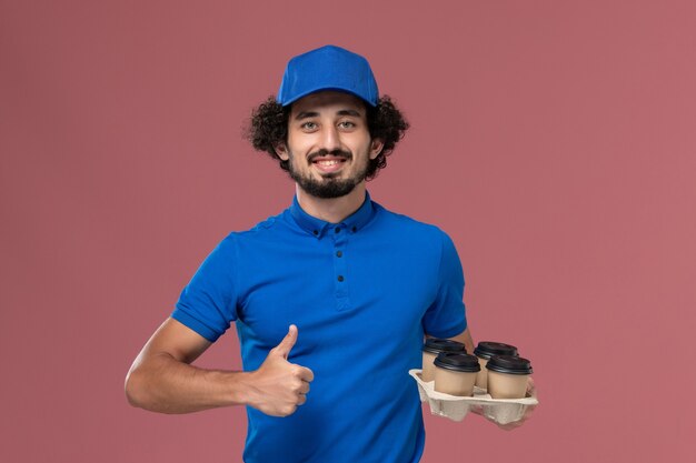 Vista frontal do mensageiro masculino com uniforme azul e boné com xícaras de café nas mãos na parede rosa claro